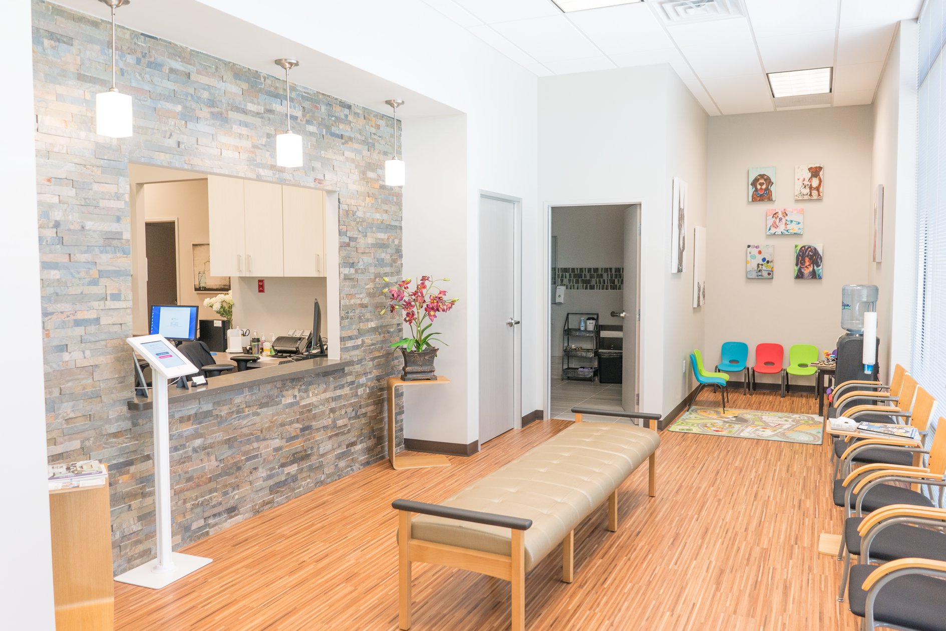 Photo of lobby and play area at University Urgent Care. Stone wall at reception area, bench in middle, chairs against the wall, and small chairs and toys in the back