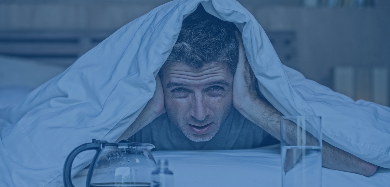 man in grey tshirt laying on bed with covers over his head with a coffee pot and glass of water - photo header for hangover treatment at university urgent care