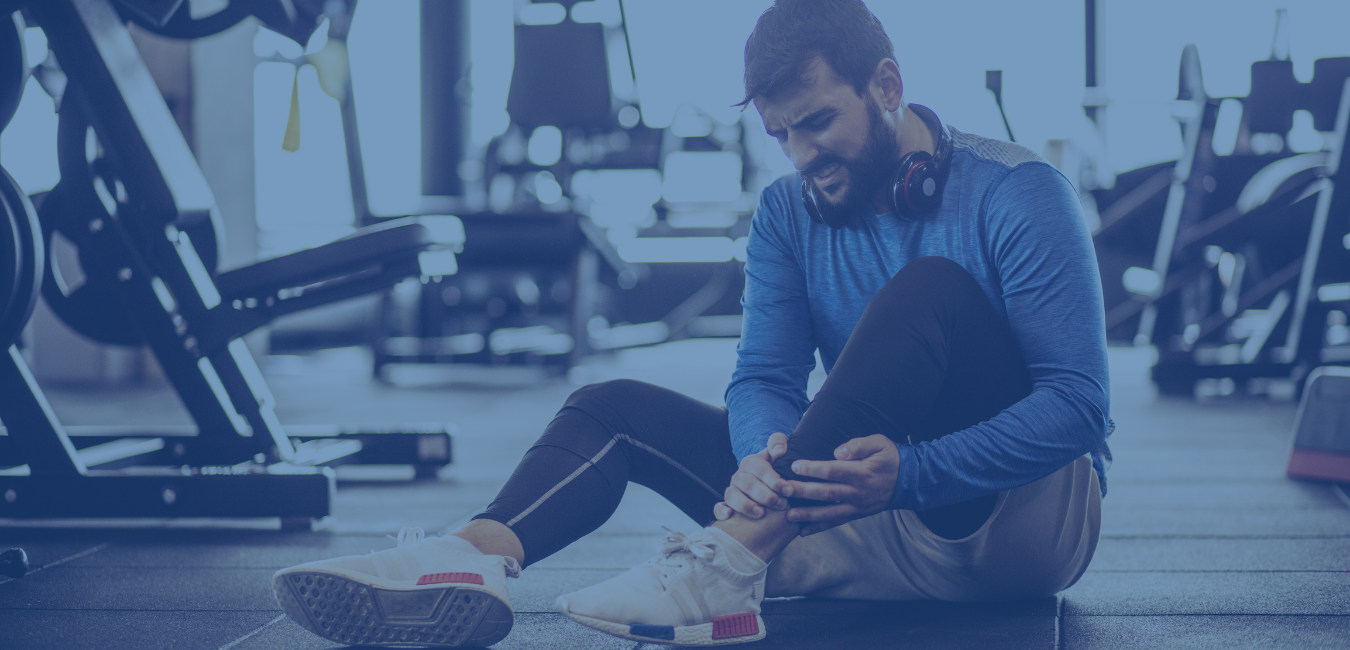 Man in blue shirt and black pants sitting on ground in gym holding ankle - header photo for sports injury page