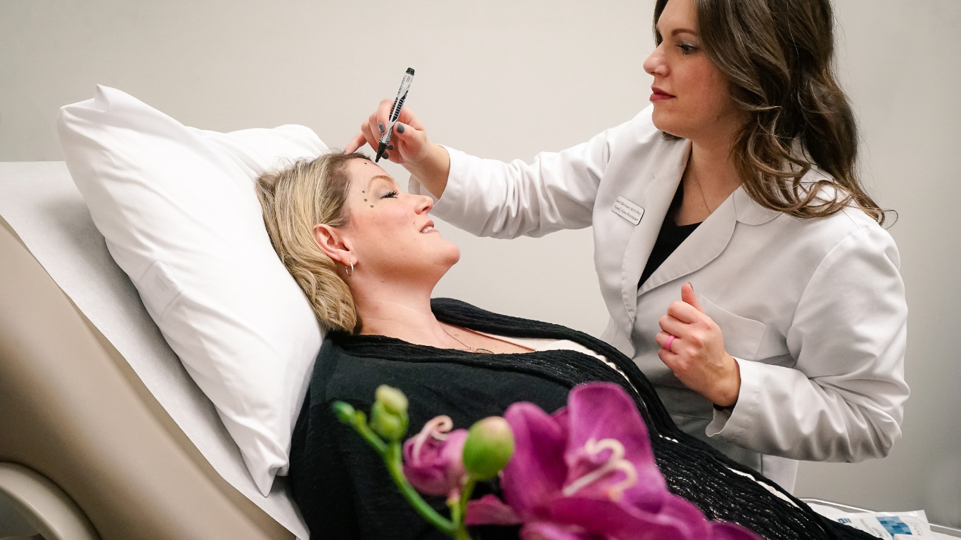 Dr injecting Botox into woman forehead. Women is wearing a black shirt and there is a pink orchid in the front of the picture